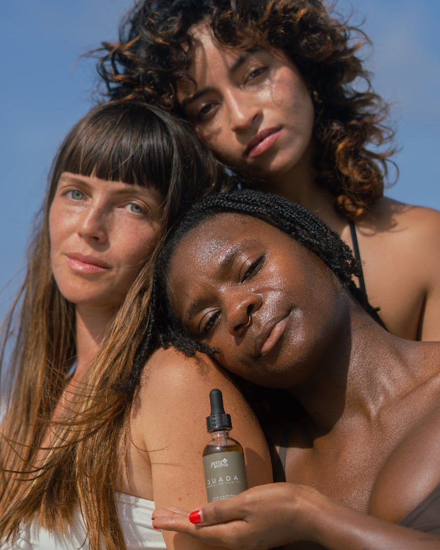 Three women standing together with a bottle of Guada nourishing hair oil against a clear blue sky.