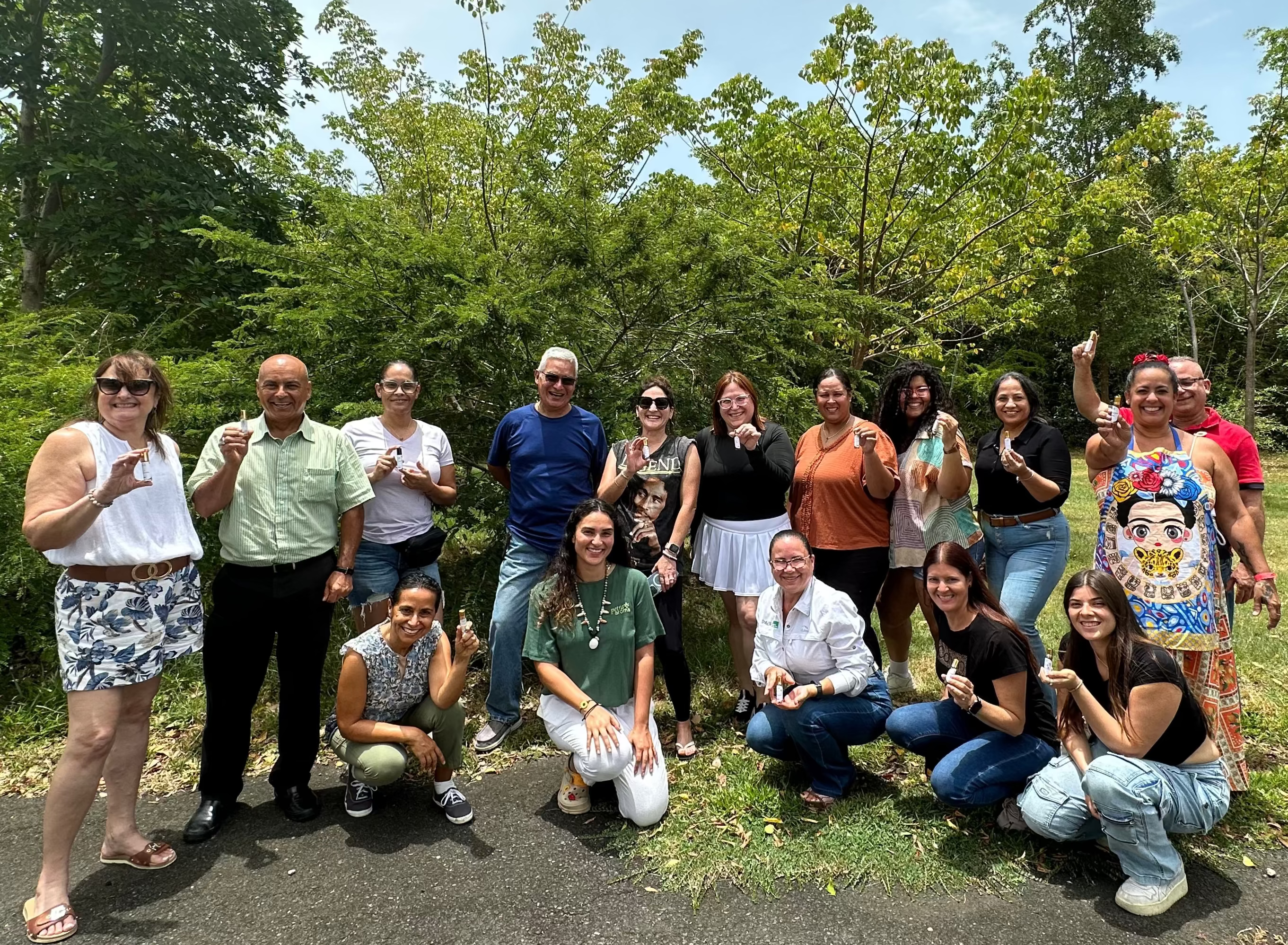 A group of about eighteen people pose outdoors standing and kneeling in front of lush green trees. They are smiling and holding up bottles of GUADA nourishing hair oil. as they gather on a paved path and grassy area. The group includes men and women of different ages, dressed in casual summer clothing such as shorts, jeans, T-shirts, and dresses. The mood is cheerful and celebratory, with natural light and greenery filling the background.
