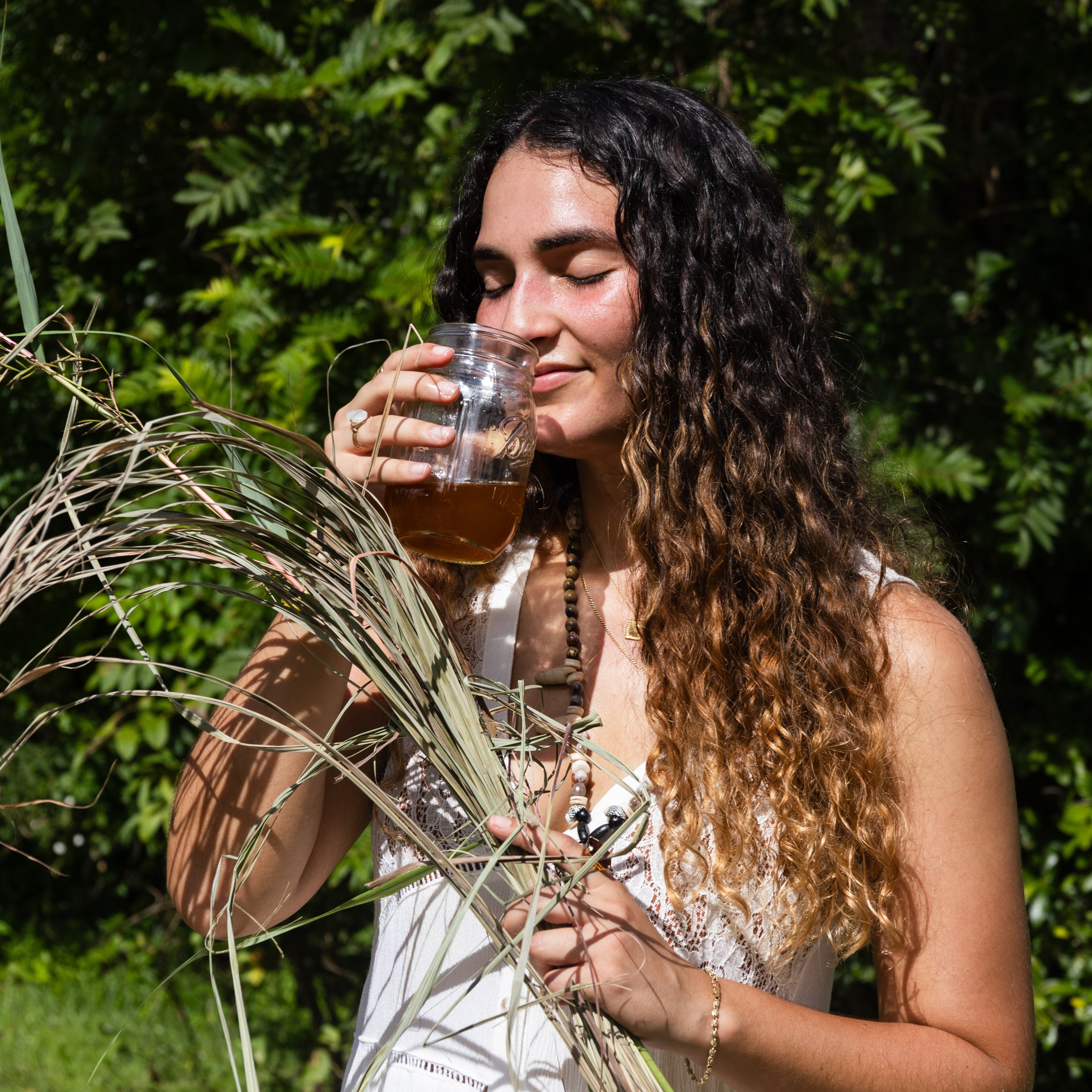 A woman with long curly brown hair stands outdoors in front of green foliage, holding a glass of amber liquid and dried grasses in the foreground. She is wearing a white sleeveless dress.